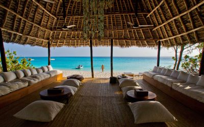 Defined Tanzania Safari A thatched-roof open-air lounge with cushioned seating overlooks a sandy beach and turquoise ocean. A person stands at the shore, and a boat floats on the water in the distance.